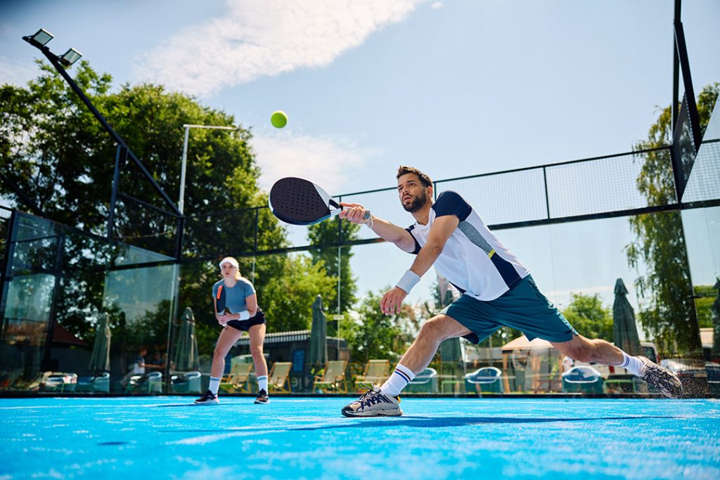 Mand er ved at modtage bolden i padeltennis imens den kvindelige medspiller afventer i baggrunden på den blå bane der er lavet af sand til padelbaner fra Dansk Kvarts Industri