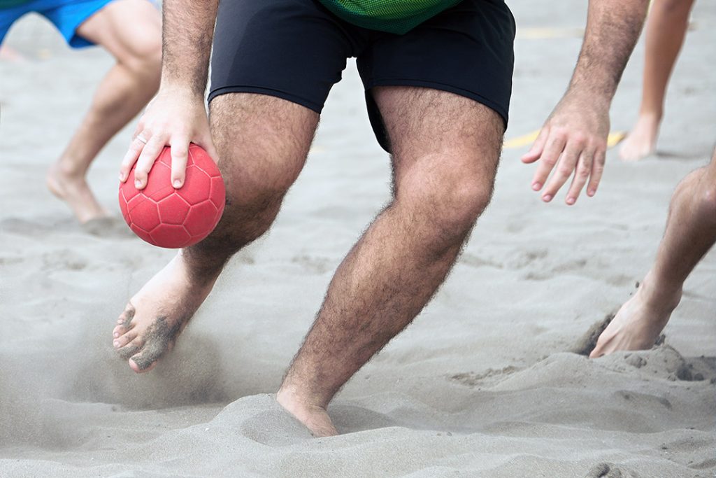 Underkrop af mand i shorts med rød håndbold i den ene hånd og som spiller strandhåndbold på strandhåndboldbane med sand til strandhåndboldbaner fra Dansk Kvarts Industri